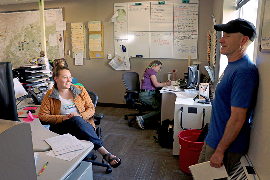 Photo of Dispatcher Carsissa Chadwick of Wenachee, Wash. talks to BLM AFS Upper Yukon Zone Duty Officer Jay Wattenbarger about a fire he is managing on June 28. In back, Terry Gallagher from Wisconsin works as a Situation Unit Leader (SITL). A SITL is responsible for collecting and organizing incident status and information and evaluating, analyzing, and displaying that information used by firefighter personnel and dispatchers.