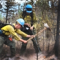 A firefighter digs up a hot spot with a pulaski while another firefighter sprays it with a hose from a portable water backpack.