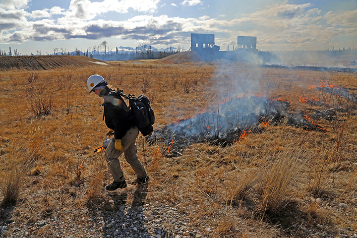 A firefighter uses a drip torch to burn dead, yellow grass in front of two military targets.