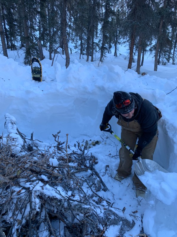 A man stands with a shovel in a snowy area with a partially dug pit.