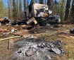 This photograph shows a pile of ashes and burned material in front of a burned camper trailer, van and other material stored outdoors.