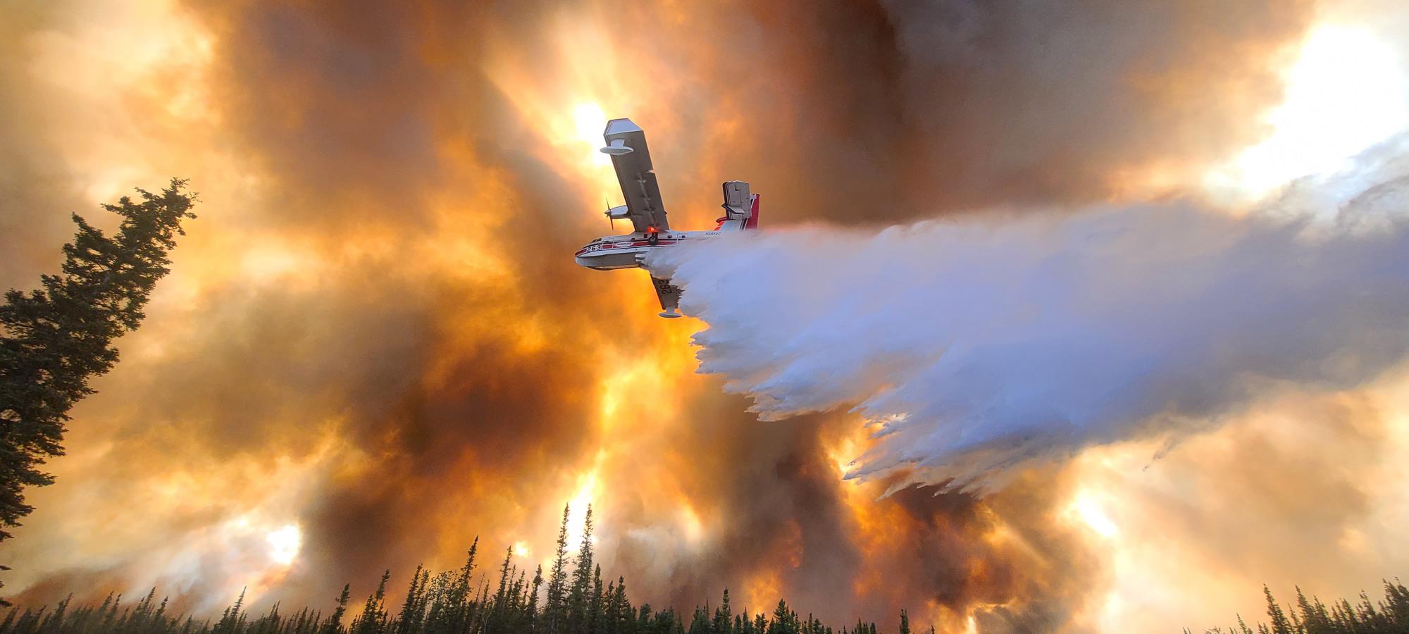 A large aircraft drops water on a fire while the sky is awash with red smoke.