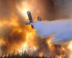 A large aircraft drops water on a fire while the sky is awash with red smoke.