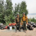 Two firefighters are standing in front of a pile of their equipment and personal gear, waiting to load into a boat. They are on a sandy beach near the Tanana River.