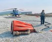 Nearest to the camera is an orange "bucket" used to hold hundreds of gallons of water that is dropped on the fire ground. On the right side of the pic are two wildland personnel, and in the back left of the photo is a stationary helicopter black in color,