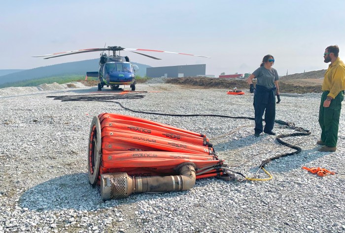Nearest to the camera is an orange "bucket" used to hold hundreds of gallons of water that is dropped on the fire ground. On the right side of the pic are two wildland personnel, and in the back left of the photo is a stationary helicopter black in color, 