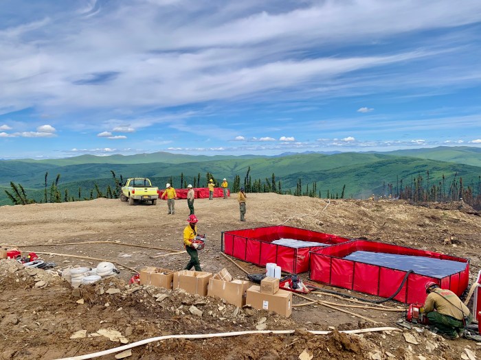 Picture of two red fold-a-tanks holding several thousands gallons of water. Boxes and several firefighers in background, looking out over vast landscape as they are on top of a ridge for the Little Chena River fire.  