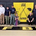A group of logistical support staff stand in a school gym in Aniak, Alaska. The school's mascot is displayed in the center.