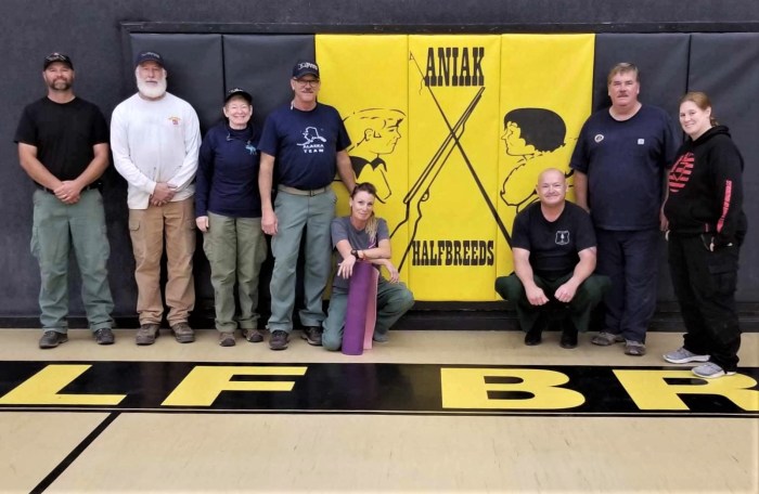 A group of logistical support staff stand in a school gym in Aniak, Alaska. The school's mascot is displayed in the center.