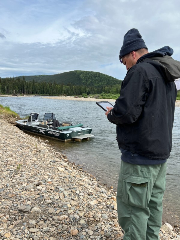 A man in black jacket and green pants stands on shore of the Salcha River while holding an iPad. River and boat are in the background with a cloudy, blue sky.