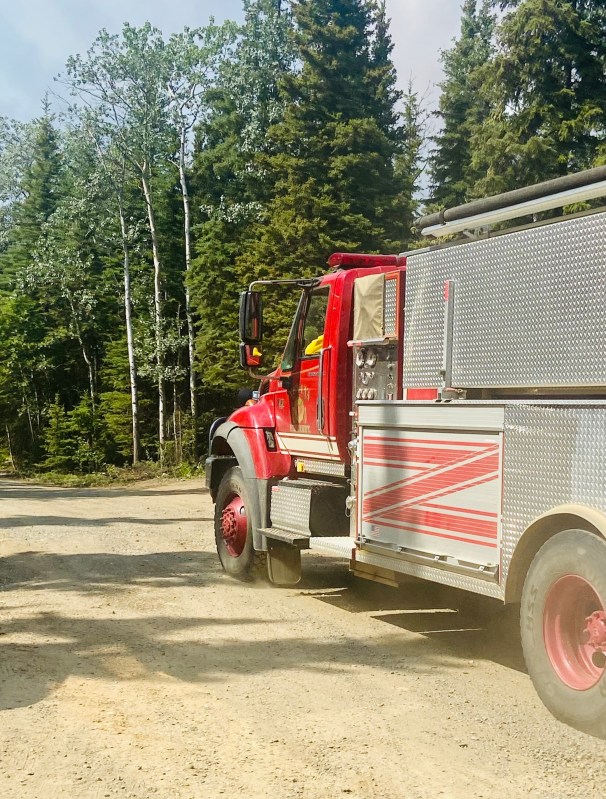 A red and white and silver water tender with Nenana Fire written on the side drives down a dirt road into an area with trees. 