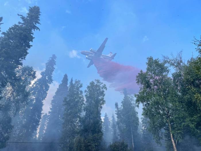 Aircraft flying above trees against a blue sky drops a red colored material known as retardant on (600) Old Ridge Trail Fire. Area has light smoke.
