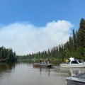 This photo is of three boats on the Goodpaster River with firefighters standing on them. There are trees along the river banks and the sky is blue with smoke billowing up in the background.