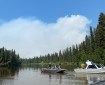 This photo is of three boats on the Goodpaster River with firefighters standing on them. There are trees along the river banks and the sky is blue with smoke billowing up in the background.