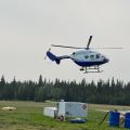Photo of a BK-117 B2 helicopter, white and blue, hovering off the ground at the helibase in Delta Junction. Fuel tanks, barrels, and various hoses sit on the grass nearby.