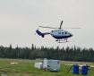 Photo of a BK-117 B2 helicopter, white and blue, hovering off the ground at the helibase in Delta Junction. Fuel tanks, barrels, and various hoses sit on the grass nearby.