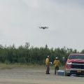 Photo of a drone in the sky with firefighters on the ground wearing yellow nomex shirts and green nomex pants. They are standing next to a red Chevrolet truck in a gravel lot. Green trees and brush in the background.