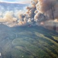 A large smoke column rises out of a fire burning through a green landscape dotted with sloughs, lakes and a river.