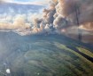 A large smoke column rises out of a fire burning through a green landscape dotted with sloughs, lakes and a river.