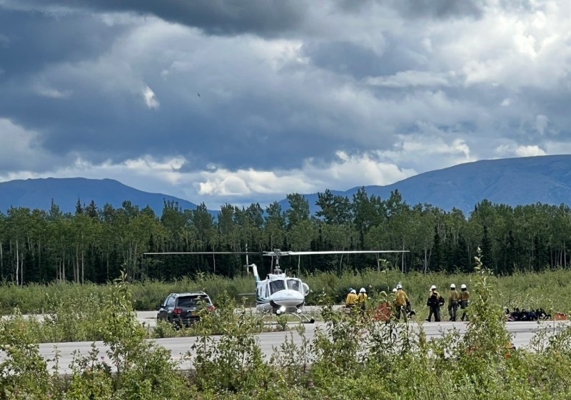 In the distance, approximately 10 firefighters in green pants, yellow shirts, and white helmets stand next to a white helicopter and wait to board for a shuttle flight to the fire.  A black car is parked on the pavement a distance away from the aircraft. Cloudy sky, mountain range and green trees in background.