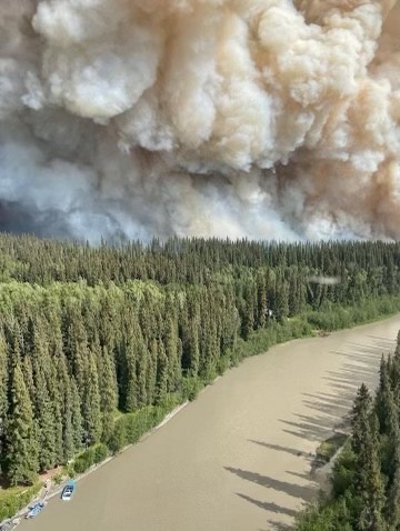 A plume of smoke raises above black spruce forest along a small river.
