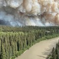 A plume of smoke raises above black spruce forest along a small river.