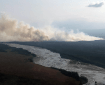 A large smoke coming up from a green landscape in back of a river.