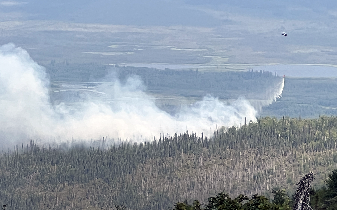 A helicopter flying with a bucket suspended beneath while dropping water on a forest fire that is producing smoke.