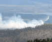 A helicopter flying with a bucket suspended beneath while dropping water on a forest fire that is producing smoke.