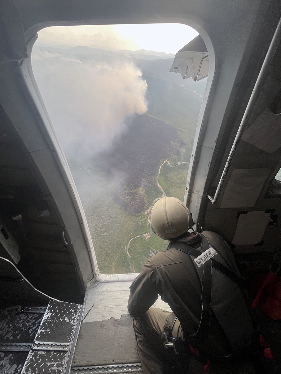 A crouching man looks out the open doorway of an airplane at a burning fire below.