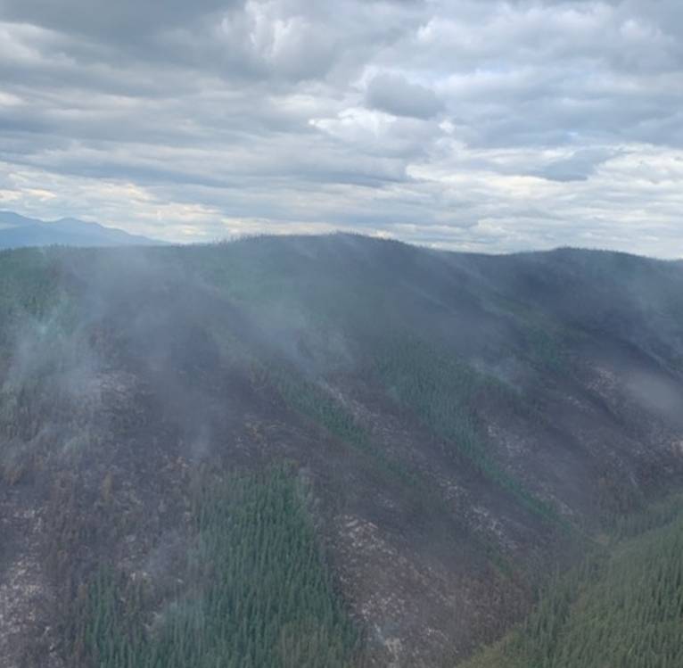Overcast skies, with wispy smoke arising from mountainside below with alternating patches of green vegetation and burned areas