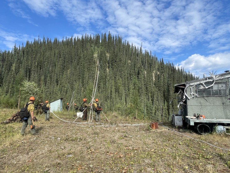 Green trees on mountain in the background. Firefigfhters erecting tall sprinkler and hose to protect building center right of image.