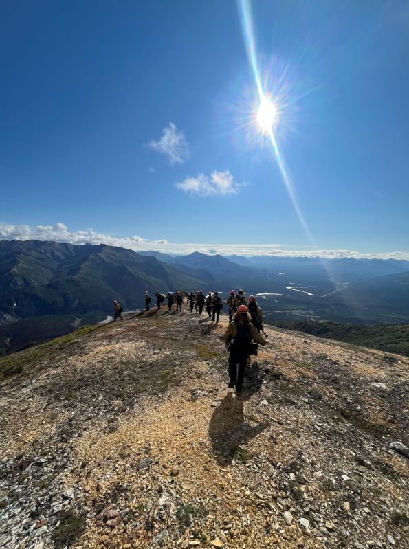 A line of firefighters hiking along a ridgeline overlooking a river and mountainside. 