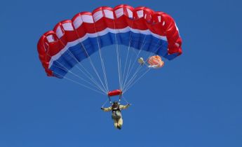 A person holds the toggles while descending to the ground underneath a red, white and blue parachute.