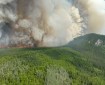 Flames and a large column of smoke rise up from a forest fire burning near a hill.