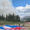 Smokejumpers on the ground pack up their gear while another firefighter descends from the air in front of a plume of smoke.