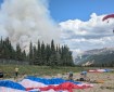Smokejumpers on the ground pack up their gear while another firefighter descends from the air in front of a plume of smoke.