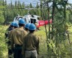 firefighters in blue hardhats and yellow shirts wait in line to board a helicopter