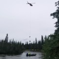 Helicopter with a slingload over a river with a boat carrying supplies.
