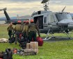 A group of wildland firefighters wearing orange hard hats line up to get on a helicopter parked on a cement pad.