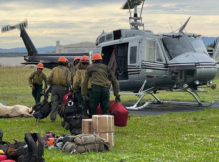 A group of wildland firefighters wearing orange hard hats line up to get on a helicopter parked on a cement pad.
