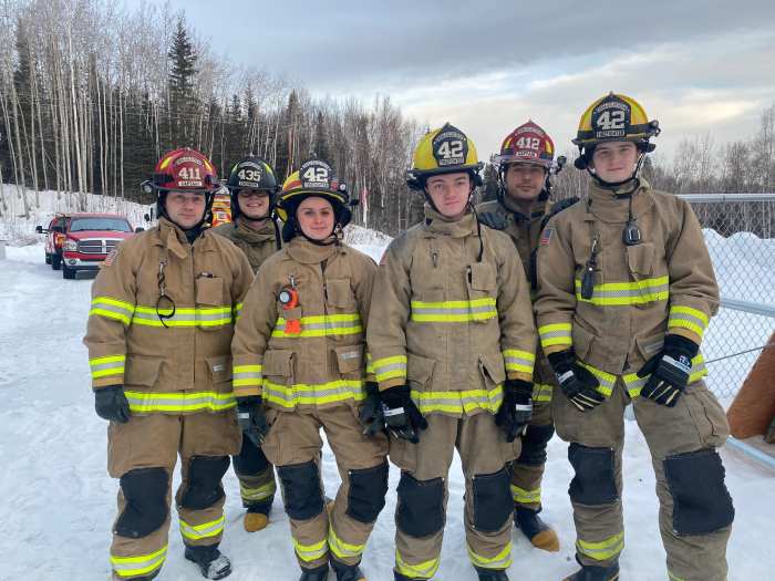 A group of 6 structure firefighters stand in a line in uniform. They are outside in a snowy setting. 