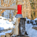 Stock photo of a hand holding an axe that is chopping firewood in the foreground. There is snow, woods, and other stacks of firewood visible in the background