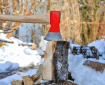 Stock photo of a hand holding an axe that is chopping firewood in the foreground. There is snow, woods, and other stacks of firewood visible in the background
