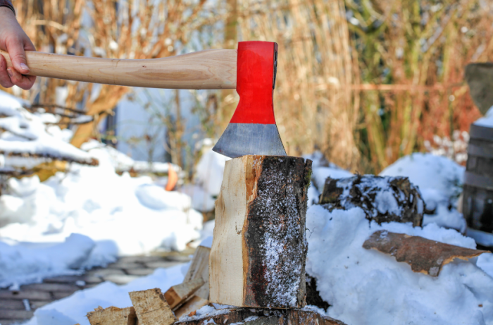 Stock photo of a hand holding an axe that is chopping firewood in the foreground. There is snow, woods, and other stacks of firewood visible in the background