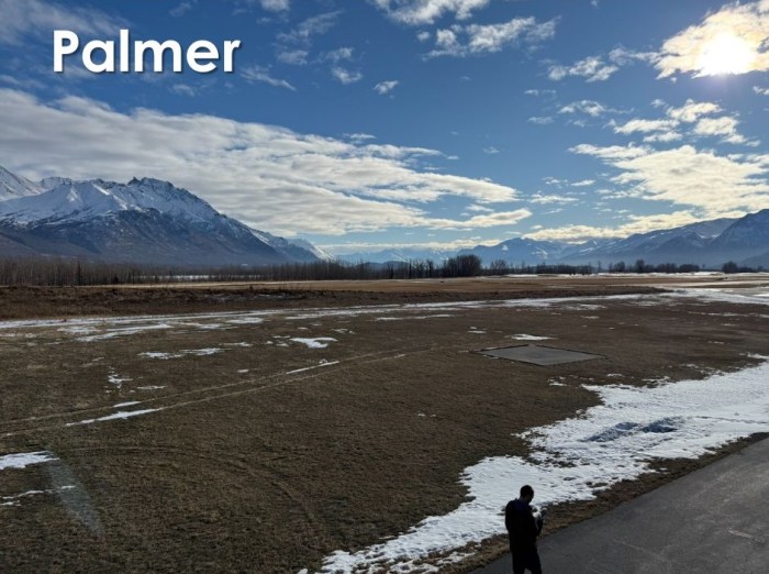 A view of dry, exposed grass surrounding the helibase and landing strip at the DOF Mat Su Area Office in Palmer. There are mountains in the background behind the helibase and a person walking in the foreground.