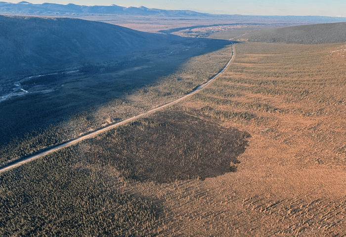 A dark burned patch of land next to a roadway with mountains in the background.