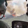 A view over the shoulder of a helicopter pilot shows fire and smoke on the ground below.