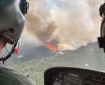 A view over the shoulder of a helicopter pilot shows fire and smoke on the ground below.