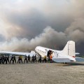 People unload gear from the left side of cargo plane as black and gray wildfire smoke billows in the background.
